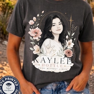 May include: A charcoal gray t-shirt featuring a graphic illustration of a person making the I love you sign in American Sign Language, surrounded by floral accents and the text Kaylee Hottle Shine Without Limits in a decorative frame.