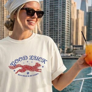 May include: Cream-coloured t-shirt featuring a red squirrel graphic and the words "Good Luck". The text "Run Wild & Be Lucky" is below the squirrel. The shirt is worn on a boat with a city skyline in the background.