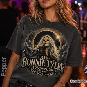 May include: A charcoal gray short-sleeve t-shirt featuring a graphic print of a singer with long hair holding a microphone. The text reads RIP BONNIE TYLER 1951-2026 TOTAL ECLIPSE TRIBUTE, surrounded by a circular sun eclipse design and clouds.