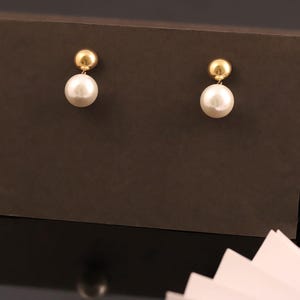 May include: A pair of gold stud earrings. Each earring has a small, round gold stud attached to a larger, white, pearlescent, spherical bead. The earrings are set against a dark brown backdrop.
