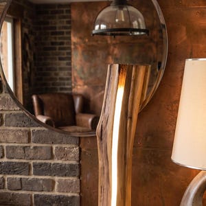 Peut inclure: Lampe de table en bois avec une bande lumineuse verticale. La lampe est en bois naturel et repose sur une surface en bois. Un miroir rond et un bol en cuivre avec des pierres sont également visibles.