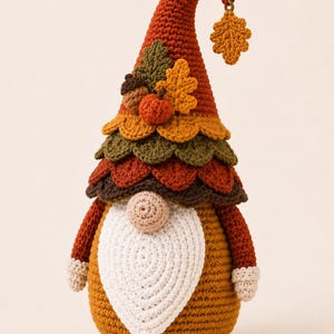 May include: A crochet gnome figurine featuring a tall, pointed rust-orange hat adorned with a small dangling leaf, a brown acorn, a tiny orange pumpkin, and several fabric leaves in shades of green and yellow. It has a white beard and beige nose.