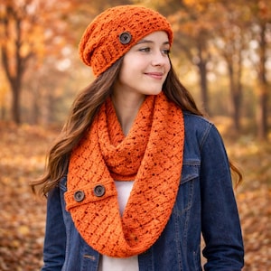 May include: An orange crochet hat and infinity scarf set with wooden buttons. The hat is a beanie style, and the scarf is a loop. The set is made of a textured, open-weave yarn. The background is a blurred autumn scene.
