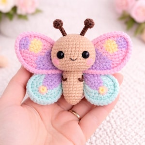 May include: A crocheted butterfly toy in pastel colors. The butterfly has a tan body, pink cheeks, and large black eyes. Its wings are pink, purple, and blue with yellow accents. The antennae are brown. The toy is held in a hand.