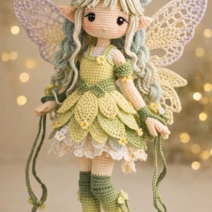 Könnte beinhalten: Eine handgefertigte Häkelfee-Puppe mit hellgrünen Haaren und zarten Gesichtszügen. Die Puppe trägt ein grünes und gelbes Blütenblattkleid, passende Armbänder und kniehohe Socken. Sie hat große, spitzenartige Flügel und hält lange grüne Schnüre.