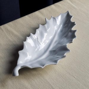 Könnte beinhalten: Eine weiße, keramikartige, blattförmige Schale mit glänzendem Finish. Die Schale hat ein detailliertes Adernmuster und einen gebogenen Stiel. Die Kanten sind gewellt, was einen dekorativen Touch verleiht. Die Schale steht auf einer beigen Oberfläche.