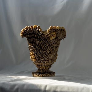 May include: A decorative vase entirely covered in a dense mosaic of small, tan, cone-shaped seashells. The vessel features a wide, flared, ruffled rim and a narrow base, with larger, smooth, rounded beige shells accenting the top and sides.