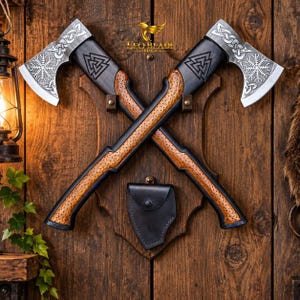 Puede incluir: Dos hachas cruzadas montadas sobre una placa de madera, con cabezas de metal plateado que presentan intrincados nudos celtas y grabados Vegvisir. Los mangos son de madera oscura con una zona de agarre texturizada en marrón claro y un símbolo Valknut.