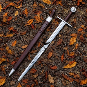 Può includere: Una spada in acciaio a doppio taglio con guardia dritta e impugnatura rivestita in pelle marrone scuro, accompagnata da un fodero in pelle marrone scuro abbinato con puntale e bocchetta in metallo argentato, posizionata su foglie secche.