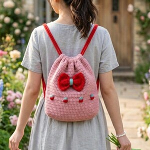以下が含まれることがあります： 赤いストラップと赤いリボンが付いたピンク色の手編みのバックパック。 バックパックには小さなイチゴの装飾と水色のリボンアクセントが付いています。 ドローストリングバッグは、庭を歩いているグレーのドレスを着た人が着用しています。