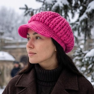 Könnte beinhalten: Eine leuchtend pinke, gestrickte Ballonmütze mit kleinem Schirm. Die Mütze ist aus dickem Garn gefertigt und hat ein strukturiertes Zopfmuster. Die Person trägt einen dunkelbraunen Mantel und einen schwarzen Rollkragenpullover. Der Hintergrund ist eine verschneite Szene im Freien.