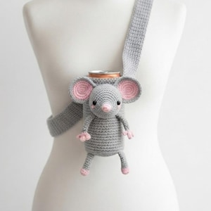 May include: A grey crocheted drink holder shaped like a mouse with pink inner ears, a pink nose, and small pink paws. The holder features a long grey strap for carrying and is designed to fit a standard beverage can securely within its body.