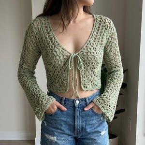 Könnte beinhalten: Ein hellgrünes Häkeltop mit offenem Gewebe, langen Ärmeln und einem Bindeband am Ausschnitt. Das Top wird mit Jeansshorts kombiniert. Das Häkeldesign erzeugt einen zarten, luftigen Look. Die Ärmel sind an den Handgelenken ausgestellt.