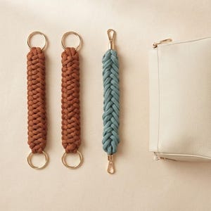 Könnte beinhalten: Zwei rostfarbene, geflochtene Kordel-Taschenriemen mit goldfarbenen Rundringen an beiden Enden, ein blaugrüner, geflochtener Kordel-Taschenriemen mit goldfarbenen Karabinerhaken an beiden Enden und eine rechteckige, cremeweiße Ledertasche.