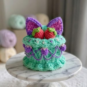 May include: A crocheted cake-shaped item in mint green, featuring a purple bow on top, two red strawberries, and small purple berries hanging from the sides. The item has a textured, ruffled base and is displayed on a white marble cake stand.