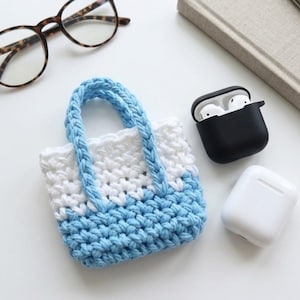 Könnte beinhalten: Eine kleine, handgefertigte Häkeltasche in Weiß und Hellblau mit passenden blauen Henkeln. Daneben eine Brille, ein schwarzes AirPod-Etui mit weißen Ohrhörern und ein weißes AirPod-Etui.