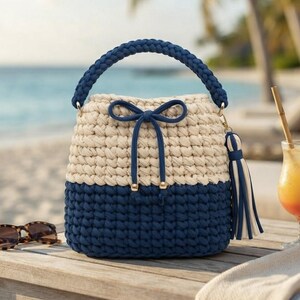 Könnte beinhalten: Eine zweifarbige Beuteltasche aus cremefarbenem und marineblauem Häkelgarn. Die Tasche verfügt über einen geflochtenen Henkel, einen Kordelzug mit Schleife und eine dekorative Quaste an der Seite. Das Design ist mit einem runden Boden strukturiert.