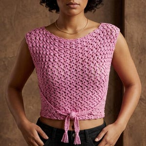 May include: A sleeveless pink crochet top featuring an intricate openwork pattern, a wide round neckline, and a tie-front detail at the hem with two dangling tassels. The garment is constructed with a textured yarn and has a cropped silhouette.