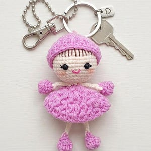 Puede incluir: Un llavero de muñeca de ganchillo hecho a mano en tonos rosa y blanco. La muñeca tiene un vestido, sombrero y zapatos rosas, con una cara sonriente. Está unido a un llavero plateado con un dije de corazón y una llave.