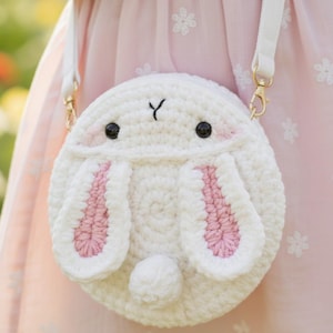 May include: A white crochet bunny-shaped crossbody bag with pink inner ears, black eyes, and a small white pom-pom tail. The bag has a white strap with gold-colored clasps. The bag is round and has a Y-shaped nose.
