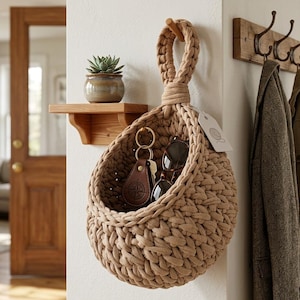 May include: A beige, woven hanging basket with a looped handle, holding keys and sunglasses. The basket is suspended from a wooden peg on a white wall. A small wooden shelf with a potted succulent is to the left.