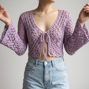May include: A light purple, open-weave crochet top with long, flared sleeves and a tie closure at the chest. The top is paired with light blue denim shorts. The crochet design features a delicate, open pattern.