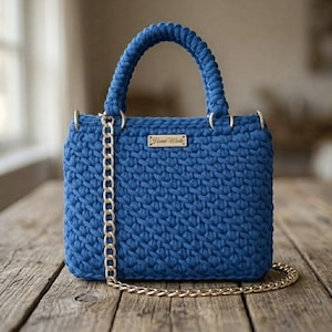 May include: A deep blue crochet handbag featuring a thick braided handle and a gold-toned metal chain shoulder strap. A small rectangular gold-colored metal tag with the text Hand Made is attached to the front center of the textured bag.