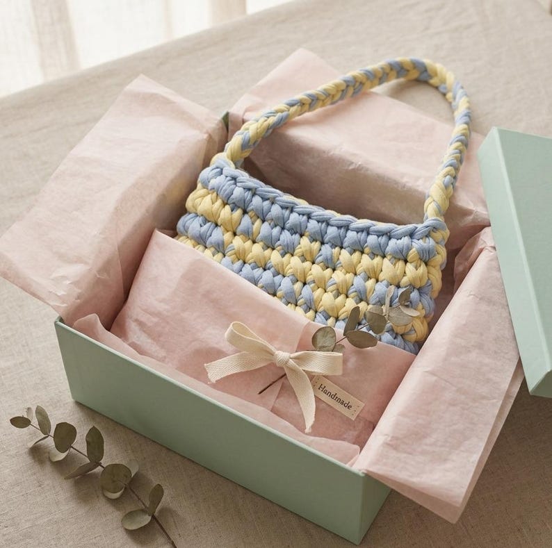 May include: A handmade crochet handbag in a gift box. The bag features alternating stripes of light blue and yellow yarn, with a matching braided handle. The gift box is a soft green color, lined with pink tissue paper, and tied with a cream-colored bow.