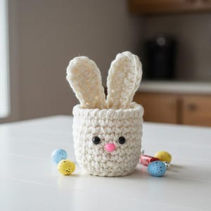 May include: A cream-colored, crocheted bunny basket with black eyes and a pink nose. The basket has two upright ears and is surrounded by colorful Easter eggs. The basket is sitting on a white surface.