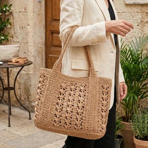 Op de afbeelding: Een bruine gehaakte tote bag met een bloemenpatroon wordt gedragen. De tas heeft twee handvatten en een rechthoekige vorm. De persoon draagt een crèmekleurige blazer en een zwarte broek. Op de achtergrond een stenen muur en potplanten.