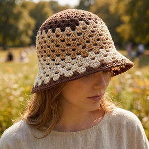 Può includere: Un cappello a secchiello all'uncinetto nei toni del marrone e del crema. Il cappello presenta un'ampia tesa e un design a trama aperta e strutturata. Il cappello è indossato all'aperto in un campo di fiori selvatici.