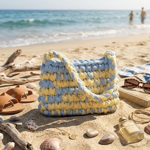 May include: A handmade, blue and yellow crochet tote bag with a braided handle sits on a sandy beach. The bag is surrounded by beach accessories, including brown sandals, sunglasses, and seashells. The ocean is visible in the background.