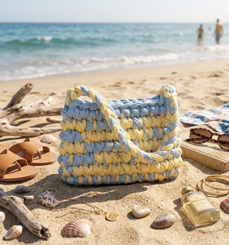May include: A handmade, blue and yellow crochet tote bag with a braided handle sits on a sandy beach. The bag is surrounded by beach accessories, including brown sandals, sunglasses, and seashells. The ocean is visible in the background.