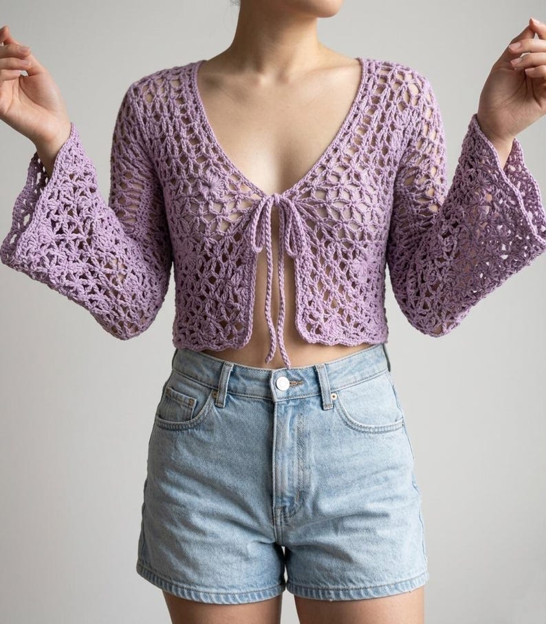 May include: A light purple, open-weave crochet top with long, flared sleeves and a tie closure at the chest. The top is paired with light blue denim shorts. The crochet design features a delicate, open pattern.