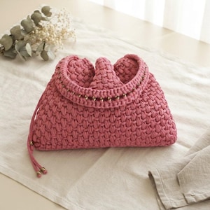 May include: A dusty rose handbag crafted from thick, woven cord in a textured pattern. The bag features a curved top opening with a decorative row of small metallic beads along the edge and two matching cord drawstrings with metallic end caps.