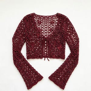 May include: A burgundy crochet top with long, flared sleeves. The open-weave design features a V-neck and a tie closure at the front. The top is made of a soft, textured yarn, perfect for layering or wearing on its own.