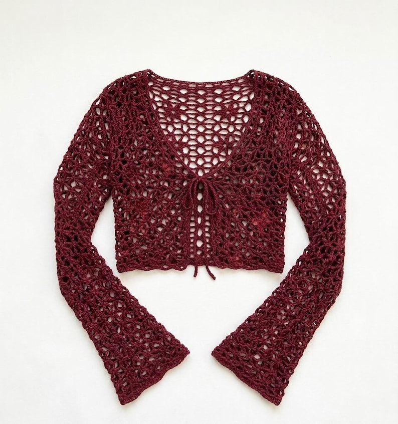 May include: A burgundy crochet top with long, flared sleeves. The open-weave design features a V-neck and a tie closure at the front. The top is made of a soft, textured yarn, perfect for layering or wearing on its own.