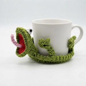 Puede incluir: Una taza de cerámica blanca envuelta en una funda de ganchillo verde con forma de rana. La rana presenta una boca muy abierta con interior rojo, una lengua rosa que sobresale y dos pequeños ojos negros y blancos. Brazos y piernas rodean la taza.