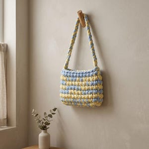 May include: A handmade, small, square-shaped crochet bag in alternating rows of light blue and yellow yarn. The bag hangs from a wooden peg on a neutral-colored wall. The strap is also crocheted in the same color pattern.