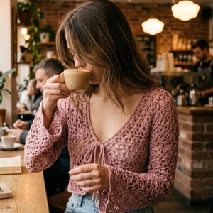 May include: A light pink crochet top with long, flared sleeves and a tie closure at the neckline. The top is paired with blue jeans. The image is taken in a cafe setting, with a person drinking from a coffee cup.