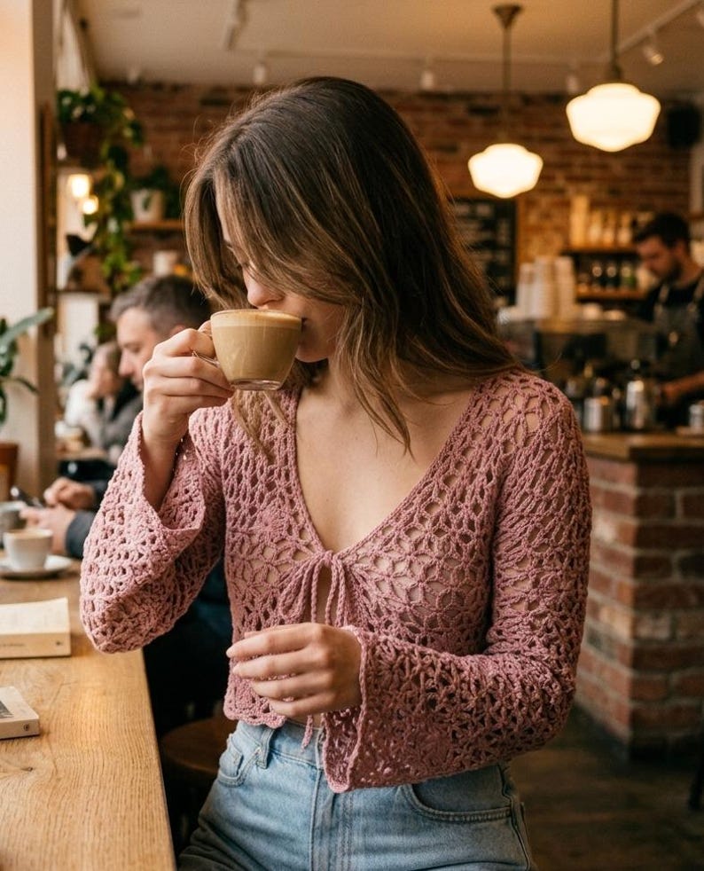 May include: A light pink crochet top with long, flared sleeves and a tie closure at the neckline. The top is paired with blue jeans. The image is taken in a cafe setting, with a person drinking from a coffee cup.