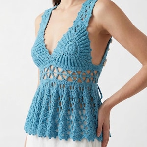 May include: A light blue crochet top with a deep V-neck and openwork detailing. The sleeveless top features a scalloped hem and a fitted bodice. The back has button closures and tie detailing. The top is made of a soft, textured yarn.