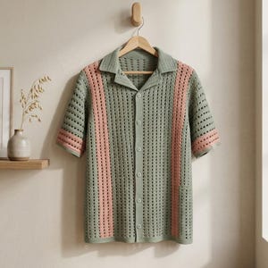 Puede incluir: Una camisa de manga corta con botones, de estilo crochet. La camisa es principalmente de color verde apagado con dos rayas verticales rosas en la parte delantera. El cuello y los pu&ntilde;os de las mangas tambi&eacute;n son rosas. La camisa cuelga de una percha de madera.