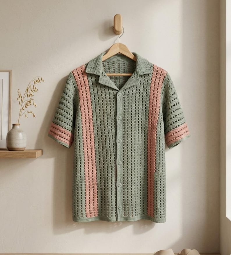 Puede incluir: Una camisa de manga corta con botones, de estilo crochet. La camisa es principalmente de color verde apagado con dos rayas verticales rosas en la parte delantera. El cuello y los pu&ntilde;os de las mangas tambi&eacute;n son rosas. La camisa cuelga de una percha de madera.