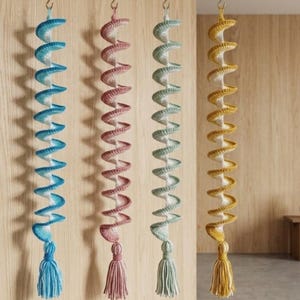 May include: Four hanging spiral crochet decorations in blue, pink, green, and yellow. Each has a tassel and a brass hook. The crochet work is detailed and the colours are soft. The decorations are suspended against a wooden background.