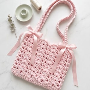 May include: A light pink crochet tote bag with two handles and decorative pink satin bows. The bag has a textured, open weave design. The bag is sitting on a white surface.