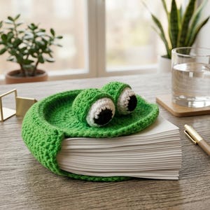 May include: A handmade green crochet frog bookmark with large white and black eyes, resting on a stack of white paper. The frog-shaped bookmark is a whimsical accessory for books and reading.