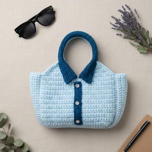 Puede incluir: Un bolso de mano de ganchillo azul claro con forma de camisa con cuello, con cuello y asa azul oscuro. El bolso tiene tres botones decorativos. Gafas de sol negras, ramitas de lavanda y un bolígrafo y cuaderno están colocados alrededor del bolso.