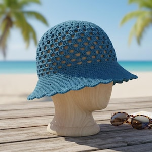 以下が含まれることがあります： 木製の頭部マネキンに、スカラップトリムが付いた青いかぎ針編みの帽子が置かれています。帽子はオープンな織り方のデザインです。 サングラスは木製の表面にあり、背景にはビーチとヤシの木が見えます。