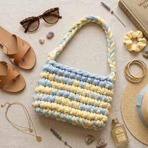 May include: A handmade, blue and yellow crochet bag with a braided handle, paired with sunglasses, sandals, jewelry, and a straw hat. The bag's design features alternating rows of blue and yellow yarn, creating a textured pattern. Other accessories include a scrunchie and a bottle of sunscreen.